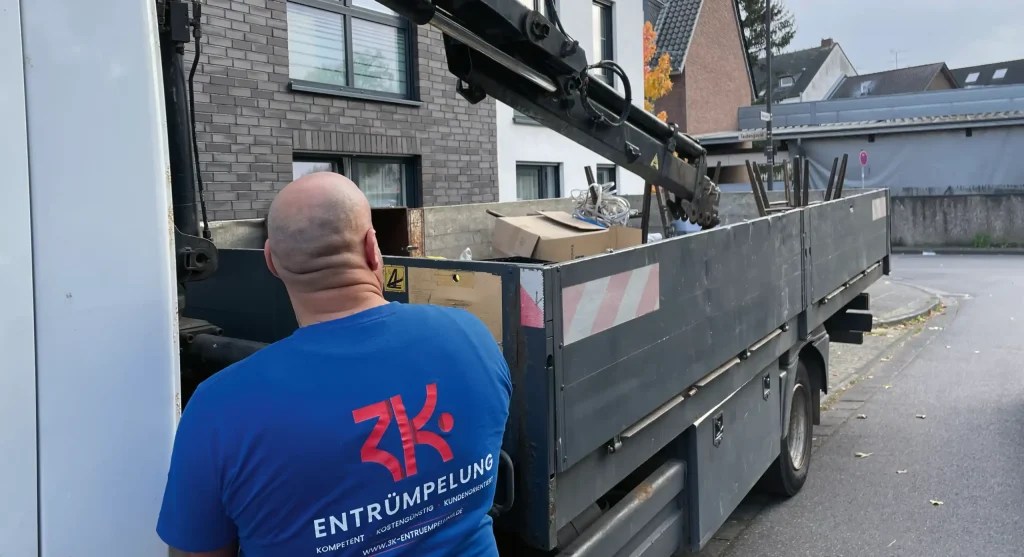 Entrümpler vor einem beladenen Transporter mit Kran, der auch für eine Entrümpelung Hennef genutzt wird. Im Hintergrund sind Wohnhäuser zu sehen.