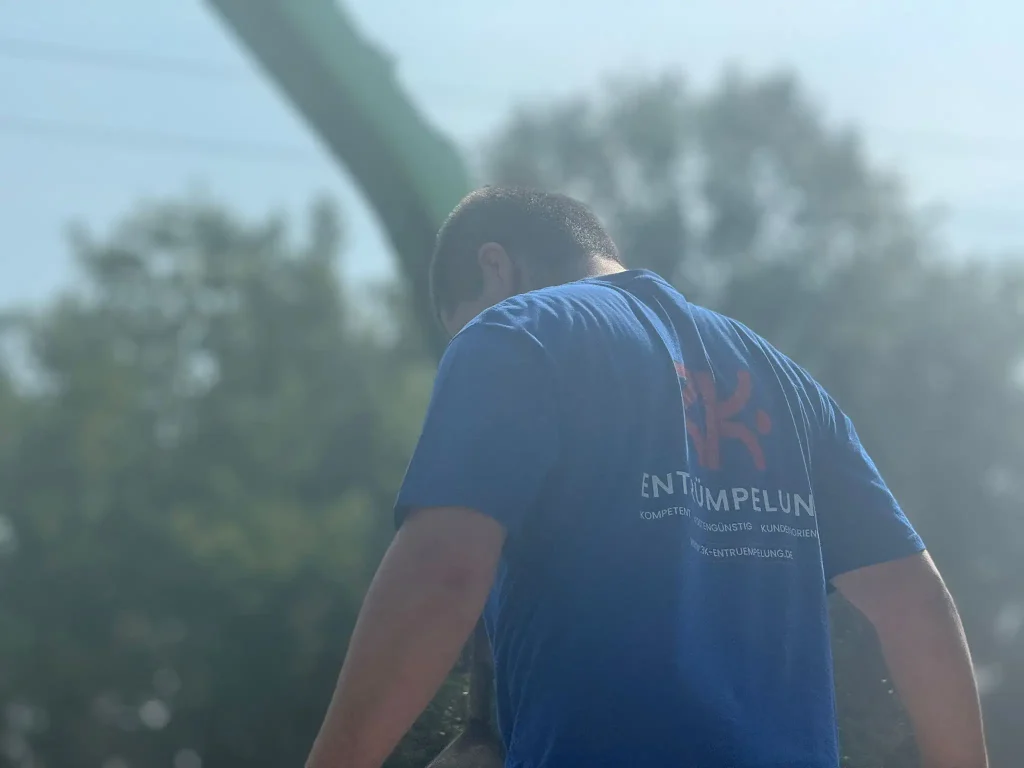 Foto - Ein Entrümpler von hinten in einem blauen T-Shirt mit dem Schriftzug "Entrümpelung" bei der Arbeit im Freien - Entrümpelung Troisdorf